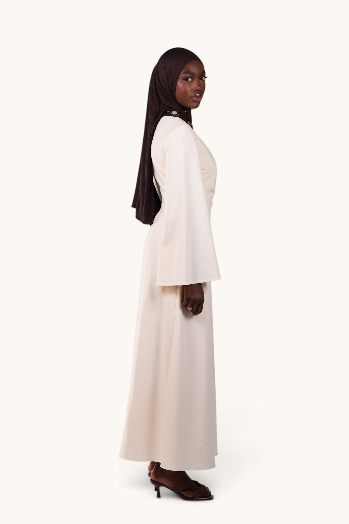 LOSYANA Valencia cream maxi dress with trumpet sleeves and gold neckline detail worn by a model in a dark hijab