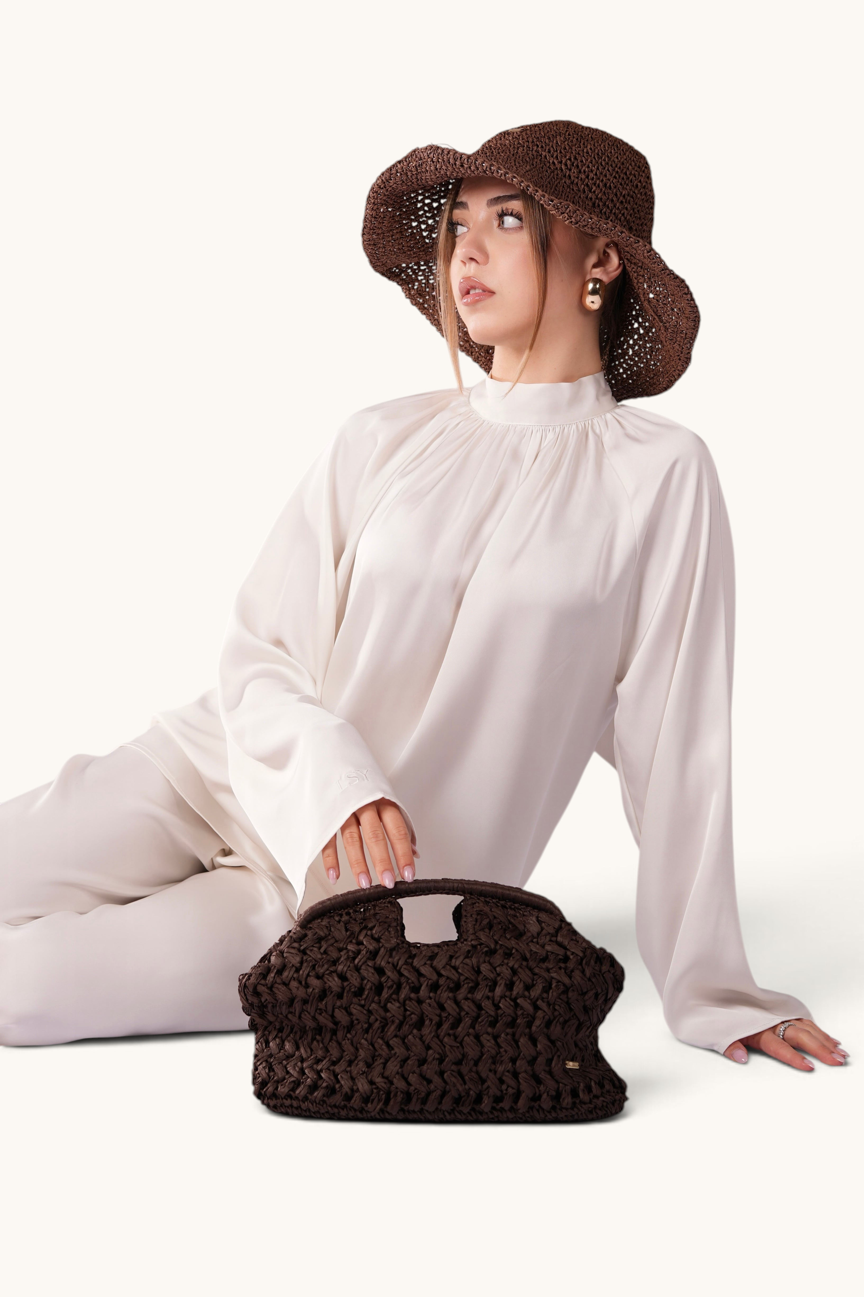 LOSYANA espresso brown woven straw handbag held by a woman wearing a matching straw hat and cream silk outfit.